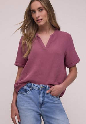 Jeune femme aux longs cheveux blonds, portant un haut ample à manches courtes rose et un jean bleu clair, debout avec une main dans la poche.