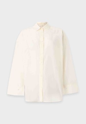 Kvinde iført en oversized cremefarvet skjorte med knapper, brede blå jeans og sorte sko, stående med den ene hånd i lommen på en ensfarvet baggrund.