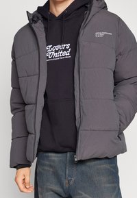 Grå polstret jakke med hætte, med en frontlynlås og synlig syning. Båret over en sort hoodie med hvid tekst. Denim jeans synlige nedenfor.