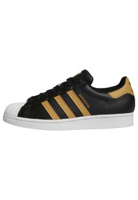 Black sneaker with three gold stripes and white rubber toe cap. Features a textured leather upper and white outsole. "Superstar" branding visible.