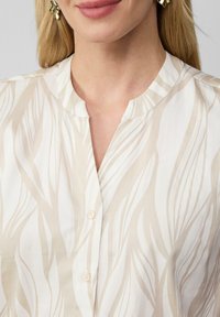 Leichte beigefarbene Bluse mit Wellenmuster, runder Kragen und kleinen Knöpfen, aus glattem Stoff mit dezentem Glanz.