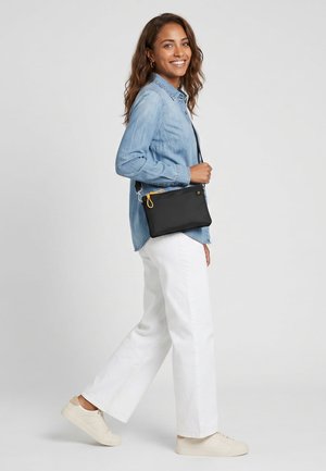Mujer sonriente caminando, con una camisa vaquera clara, pantalones blancos, zapatillas crema y llevando un bolso negro de bandolera con cremallera amarilla.