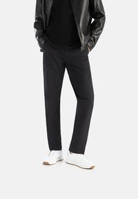 Sorte, lige bukser lavet af glat stof, parret med hvide sneakers med en tekstureret sål. Modellen bærer en sort skjorte og læderjakke.