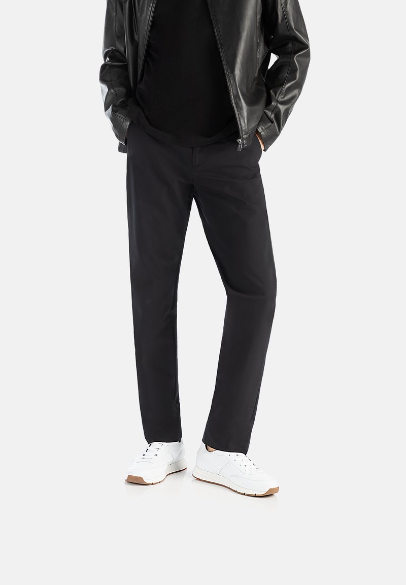 Sorte, lige bukser lavet af glat stof, parret med hvide sneakers med en tekstureret sål. Modellen bærer en sort skjorte og læderjakke.