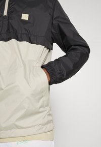 Giacca a pullover con una parte superiore nera e una parte inferiore beige chiaro. Realizzata in materiale elegante e resistente all'acqua con polsini elastici.