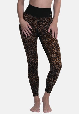 Frau trägt hoch taillierte Leggings mit Leopardenmuster und einem schwarzen Bund, steht barfuß mit den Händen auf den Oberschenkeln.