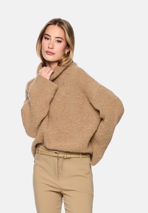 Junge Frau trägt einen hellbraunen flauschigen Pullover und beige Hosen mit einem goldenen Kettengürtel, posiert mit einer Hand nahe am Kinn.