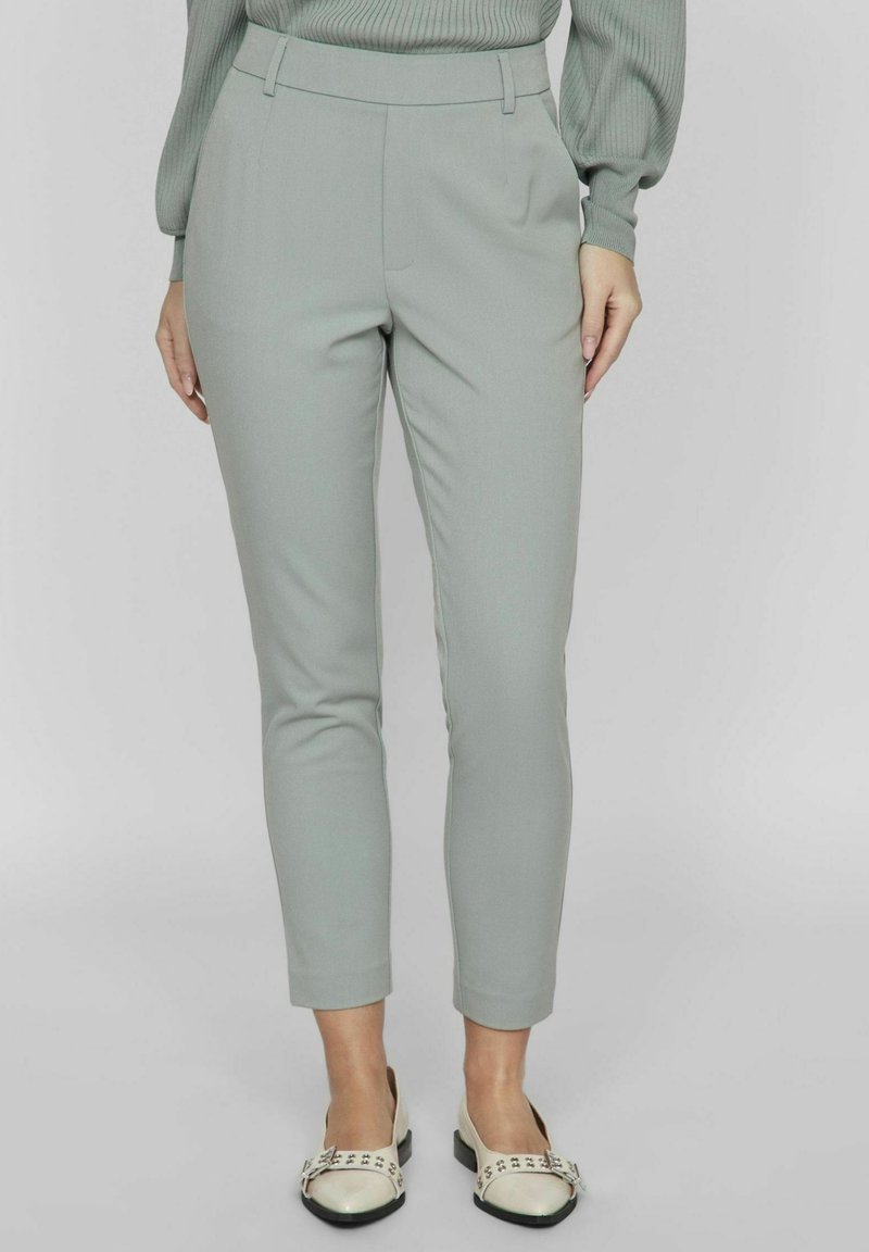 Vaaleanharmaat kapenevat housut, joissa on sileä kangas, korkea vyötärö ja sivutaskut. Yhdistettyinä kermaiseen kenkäpariin, jossa on mustia yksityiskohtia.