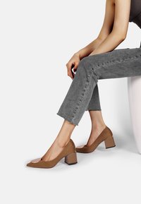Puntini alti in suede marrone con punta quadrata, caratterizzati da un accento lucido sulla tomaia e un tacco a blocco largo. Indossati con jeans grigi a vita alta.