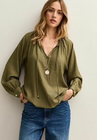 Olijfgroene, lichte, lange mouwen blouse met een V-hals, trekkoorddetail, getextureerde stof en knoopmanchetten, gecombineerd met spijkerbroek.