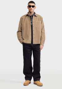 Jasnobrązowa koszula overshirt z kordurowego materiału z zatrzaskami, czarna koszulka pod spodem, czarne dżinsy i beżowe buty. Model ma także okulary przeciwsłoneczne.