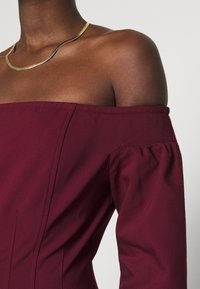 Burgunderfarbenes Off-Shoulder-Oberteil mit Puffärmeln und Nahtdetails, ergänzt durch eine glatte metallic-goldene Halskette.