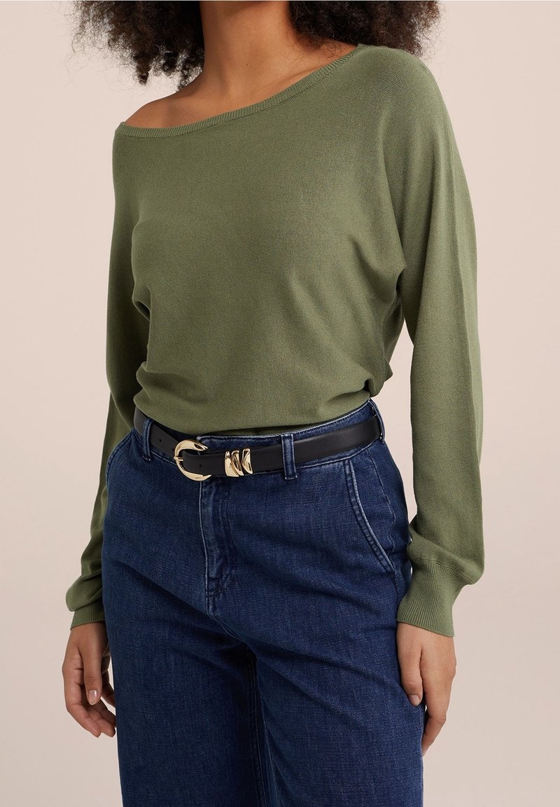 Pull vert olive à épaules dénudées associé à un jean bleu foncé et une ceinture noire avec une boucle dorée. Texture lisse et coupe décontractée.