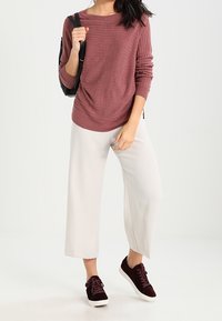 Bordeauxrotes Langarmoberteil aus strukturiertem Stoff, cremefarbene weite Hose und dunkelburgunderfarbene Wildledersneaker. Schwarzer Rucksack auf der Schulter.