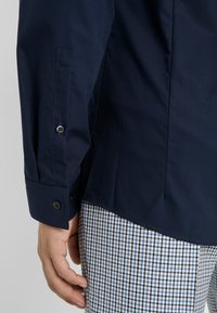 Main gauche détendue le long du corps, portant une chemise à manches longues bleu foncé et un pantalon à carreaux bleu, blanc et noir sur fond neutre.