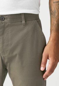 Olive green trousers in a smooth fabric with a flat front, side pockets, and a button closure, featuring subtle stitching details.