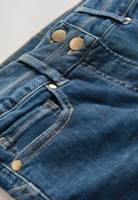 Temno modri kavbojki iz denima z zlatimi gumbki, šivanim okrasjem in klasičnim petim žepnim dizajnom, ki prikazujejo gladko teksturo.