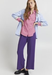 Hellblaues Blazer mit gepunkteten Manschetten, rosa Hemd und weite lila Hosen. Das Modell steht mit verschränkten Armen. Schwarze Schuhe sind sichtbar.
