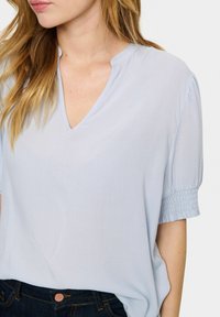 Blusa azzurro chiaro a maniche corte con scollatura a V e tessuto testurizzato; presenta dettagli arricciati sulle maniche e una vestibilità morbida.