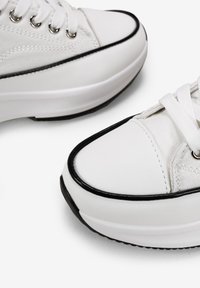 White canvas sneakers with black rubber accents, round toe, contrasting stitching, and metal eyelets for laces. Smooth texture, flat sole.