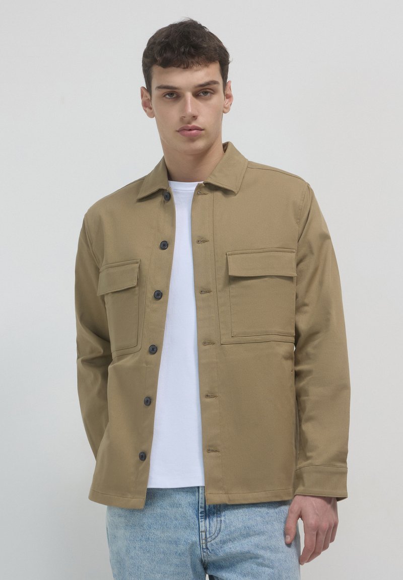 Junger Mann in einer beigen Knopfjacke über einem weißen Hemd und hellblauen Jeans, der vor einem einfarbigen hellgrauen Hintergrund steht.