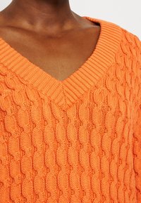Sweter o splocie warkoczowym w jaskrawym pomarańczowym kolorze, z dekoltem w kształcie V i ściśle przylegającymi wykończeniami. Posiada teksturowany wzór z równomiernymi, grubymi ściegami.