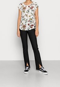 Kvinde iført en hvid blomstret kortærmet bluse, sorte tætsiddende bukser med frontslids og sort-hvide sneakers, stående på et lyst gulv.