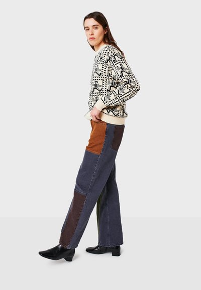 Maglione a maglia con motivo geometrico in nero e crema; pantaloni in denim con un patchwork multicolore in marrone, verde e blu; stivaletti neri alla caviglia.