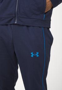 Marine idretts bukser med blå accenter og Under Armour-logo. Har en myk tekstur og glidelåslommer for funksjonalitet.