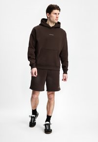 Brun hettegenser og matchende shorts-sett, med en frontlomme og logo detaljer, laget av teksturert stoff, sammen med svarte sko.