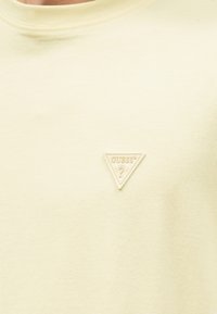 Tissu couleur crème avec un petit patch triangulaire du logo GUESS comportant un point d'interrogation sur la partie supérieure de la poitrine.