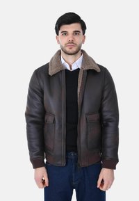 Uomo che indossa una giacca in pelle shearling marrone scuro sopra un maglione nero e una camicia bianca con colletto, in piedi su uno sfondo bianco uniforme.