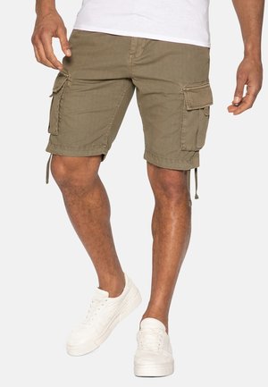 Homme portant un short cargo vert olive et des baskets blanches, debout devant un fond clair uni.