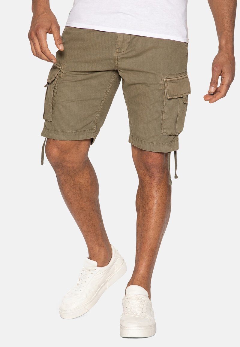 Homme portant un short cargo vert olive et des baskets blanches, debout devant un fond clair uni.