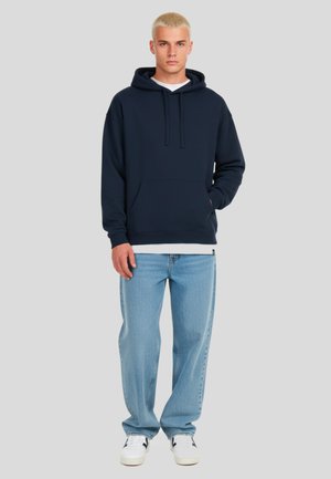 Felpa navy con tasca a marsupio, cappuccio regolabile e polsini a costine, abbinata a jeans dritti azzurri e sneakers bianche.