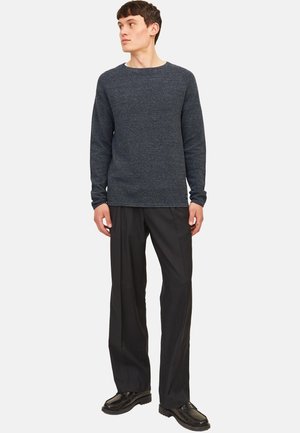 Jeune homme debout, portant un pull à manches longues gris foncé, un pantalon noir ample et des chaussures en cuir noires, regardant sur le côté.