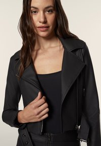 Giacca da moto in pelle sintetica nera con zip asimmetrica, rever ampi e design aderente. Presenta tasche con zip e una texture liscia.