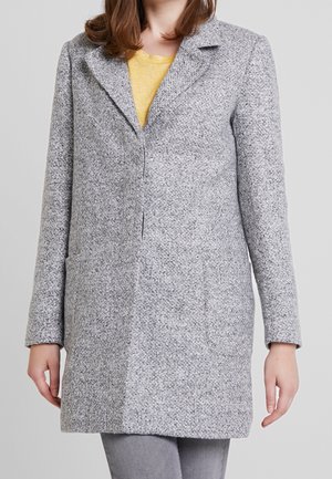 Femme portant un manteau gris clair texturé sur un top jaune et un pantalon gris clair, montrée du cou à la mi-cuisse sur un fond uni.