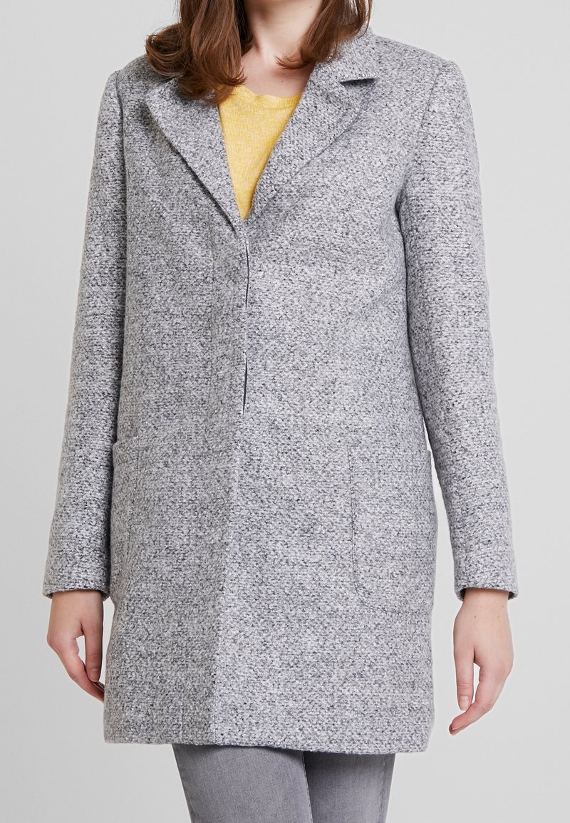 Femme portant un manteau gris clair texturé sur un top jaune et un pantalon gris clair, montrée du cou à la mi-cuisse sur un fond uni.