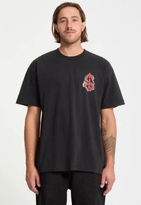 Black cotton t-shirt with short sleeves. Features a red and white graphic design on the left chest area. Relaxed fit and crew neckline.