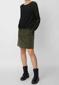 Schwarze Bluse mit langen Ärmeln und gerafftem Saum, kombiniert mit einem knielangen olivgrünen Cordrock und schwarzen Schnürkampfstiefeln.