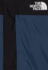 Chaqueta azul y negra con una tela suave y brillante de textura en cuadrícula, con costuras doradas y un logotipo blanco prominente en la parte superior.