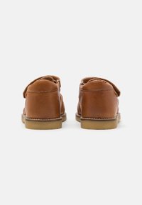 Scarpe in pelle marrone con superficie liscia, tacco arrotondato e cinturino in velcro. Presentano una suola in gomma color sabbia e dettagli di cucitura minimi.
