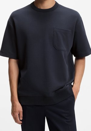 Homme portant un t-shirt uni bleu marine foncé à manches courtes avec une poche poitrine, debout avec une main dans la poche, sur fond blanc.