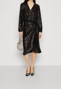 Svart paljettomslagsklänning med lång ärm, med en V-ringning och rynkad midja. Kombinerad med utsmyckade svarta klackar och en silverväska.