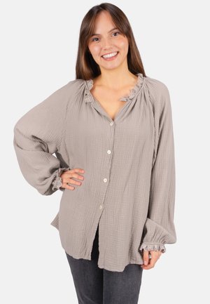 Femme souriante portant un chemisier beige ample à boutons avec un col et des poignets à volants, posant avec une main sur la hanche sur un fond blanc.