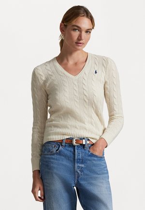Crème kabelgebreide trui met V-hals en lange mouwen, voorzien van een klein blauw logo op de borst, gecombineerd met lichtblauwe jeans en een bruine riem.