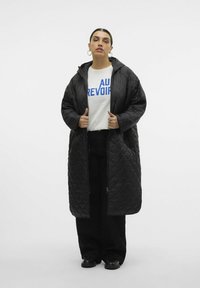 Zwarte gewatteerde lange jas met capuchon, voorrits en zijzakken, gedragen over een wit shirt met blauwe tekst. Dikke stof, losse pasvorm.