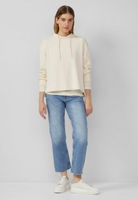 Cremfarbener Kapuzenpullover mit Kordeln, über einem hell gestreepten Oberteil getragen, kombiniert mit blauen geraden Jeans und weißen Plateausneakern.