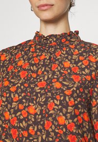 Blusa floreale con un colletto alto arricciato, tessuto marrone scuro con fiori rossi e gialli e motivi di foglie verdi.